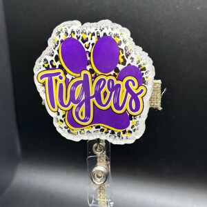 May include: A purple and gold glitter paw print with the word "Tigers" written in a purple script. The paw print is attached to a clear plastic retractable badge holder with a brown cord.