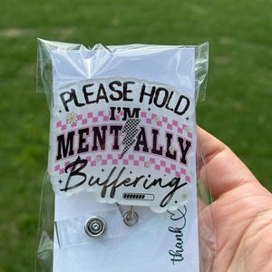 Peut inclure: Un badge transparent avec le texte "PLEASE HOLD I'M MENTALLY Buffering" en noir et rose. Le design comprend un motif à carreaux roses et blancs, un éclair et une barre de chargement. Le badge est emballé dans un sac en plastique transparent.