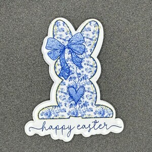 May include: A blue and white floral patterned Easter bunny with a blue bow and a blue heart on its chest. The bunny is surrounded by a white outline and the text "happy easter" is written below it.