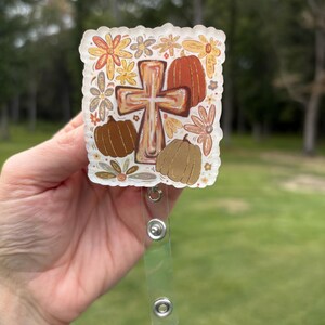 May include: A clear, square badge reel featuring a cross, pumpkins, and floral designs in shades of brown, orange, and yellow. The badge reel has a clear plastic strap and a metal clip for attaching to clothing or accessories.