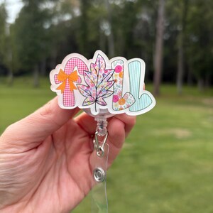 May include: A badge reel with the word "FALL" in colorful, patterned letters. The "F" is a candy cane with an orange bow, the "A" is a leaf, and the "L"s are floral and striped. The reel has a clear plastic strap.