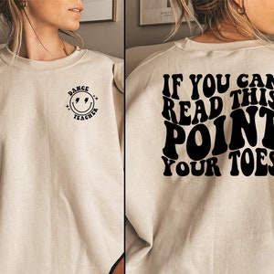 May include: A beige sweatshirt with a black smiley face and the text "Dance Teacher" on the front. The back of the sweatshirt has the text "If you can read this point your toes" in a retro font.