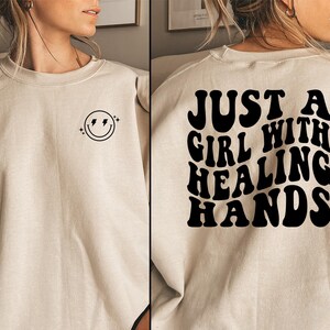 May include: A beige sweatshirt with a smiley face and lightning bolt design on the front. The back of the sweatshirt features the text "JUST A GIRL WITH HEALING HANDS" in black, retro-style lettering.