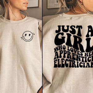 May include: A beige sweatshirt with a smiley face with lightning bolts on the left side and the text "Just a girl who loves her apprentice electrician" on the right side.