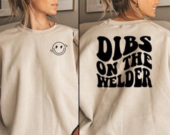 May include: A beige sweatshirt with a black smiley face with lightning bolts on the front and the text "DIBS ON THE WELDER" in a retro font on the back.