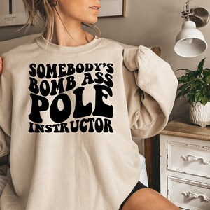 May include: A beige sweatshirt with the text "Somebody's Bomb Ass Pole Instructor" in black retro font.