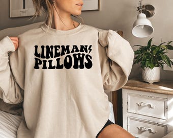 May include: A beige sweatshirt with the text "LINEMAN/PILLOWS" in black retro-style lettering.