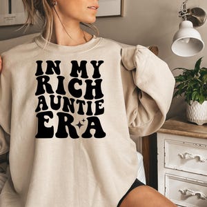 May include: A beige crewneck sweatshirt featuring the black text "IN MY RICH AUNTIE ERA" printed on the front in a bold, wavy font. The design includes two small four-pointed star icons positioned between the words "ERA" and "A".