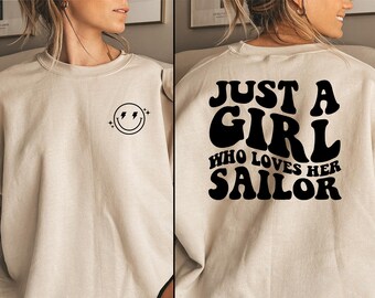 May include: Beige sweatshirt with a smiley face graphic with lightning bolts on the front. The back of the sweatshirt features the text "JUST A GIRL WHO LOVES HER SAILOR" in black block letters. A casual, comfortable top.
