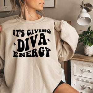 May include: A beige sweatshirt with black text that reads "It's Giving Diva Energy".