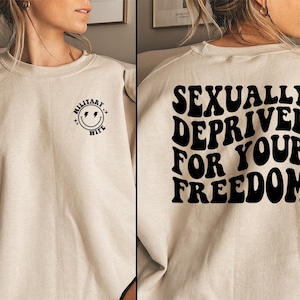 May include: A beige sweatshirt with black text on the front and back. The front features a smiley face with lightning bolts and the words "MILITARY WIFE". The back says "SEXUALLY DEPRIVED FOR YOUR FREEDOM" in a retro font.