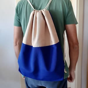 以下が含まれることがあります： 白の紐が付いたベージュとブルーの巾着袋。人が背負っています。