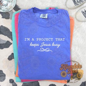 May include: A stack of colorful t-shirts in shades of blue, pink, and orange. The top shirt is blue and features the text "I'M A PROJECT THAT keeps Jesus busy" in white. The shirts are folded and stacked, with a logo in the bottom right corner.