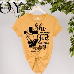 Op de afbeelding: Een mosterdgeel T-shirt met een zwart grafisch ontwerp van gevouwen handen met een kruis en de tekst "She who kneels before God can stand before anyone Romans 8:31"