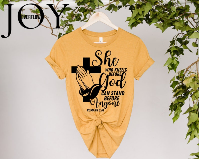 Op de afbeelding: Een mosterdgeel T-shirt met een zwart grafisch ontwerp van gevouwen handen met een kruis en de tekst "She who kneels before God can stand before anyone Romans 8:31"