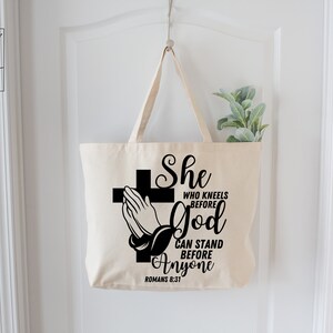 Op de afbeelding: Een witte canvas tote bag met zwarte tekst en een afbeelding van een biddend hand voor een kruis. De tekst luidt "She who kneels before God can stand before anyone Romans 8:31".