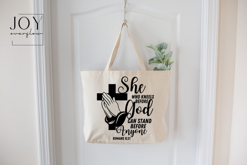 Op de afbeelding: Een witte canvas tote bag met zwarte tekst en een afbeelding van een biddend hand voor een kruis. De tekst luidt "She who kneels before God can stand before anyone Romans 8:31".
