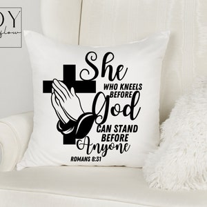 Op de afbeelding: Een wit kussen met zwarte tekst die luidt "She who kneels before God can stand before anyone Romans 8:31". De tekst is in een decoratief lettertype en bevat een kruis met een biddend hand.
