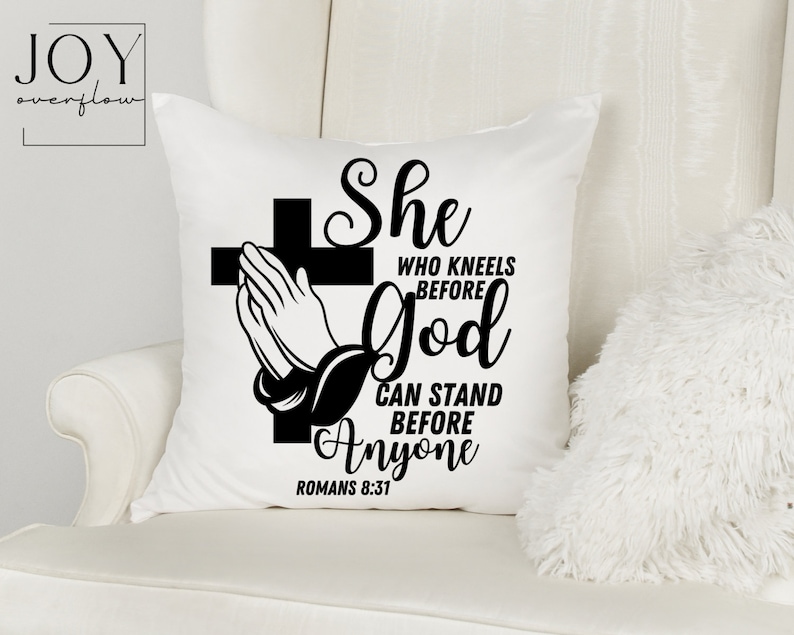 Op de afbeelding: Een wit kussen met zwarte tekst die luidt "She who kneels before God can stand before anyone Romans 8:31". De tekst is in een decoratief lettertype en bevat een kruis met een biddend hand.