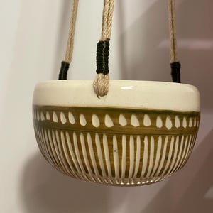 Peut inclure: Un pot de fleurs suspendu en céramique avec un dessus crème et un fond marron à motifs. Le pot est suspendu par trois cordes beiges avec des accents noirs. Le design présente des lignes verticales et des motifs pointillés.