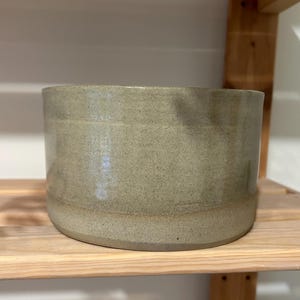 May include: A cylindrical ceramic bowl with a speckled light sage green glaze. The vessel features a smooth, uniform exterior surface with subtle tonal variations and a flat base, showcasing a simple, functional design for everyday use.