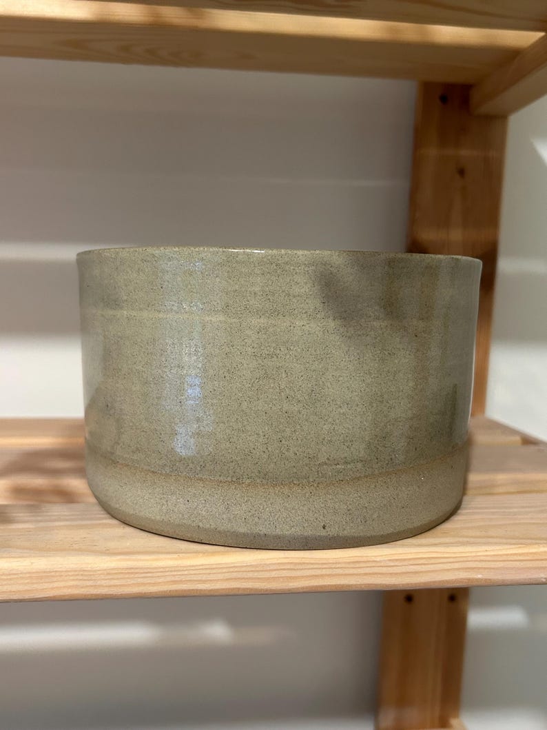 May include: A cylindrical ceramic bowl with a speckled light sage green glaze. The vessel features a smooth, uniform exterior surface with subtle tonal variations and a flat base, showcasing a simple, functional design for everyday use.