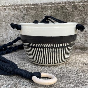 Peut inclure: Un pot de fleurs suspendu en céramique blanche avec un motif géométrique noir. Le pot a un support en macramé noir avec un anneau en bois. Le pot est conçu pour une utilisation intérieure ou extérieure et est une façon élégante d'exposer les plantes.