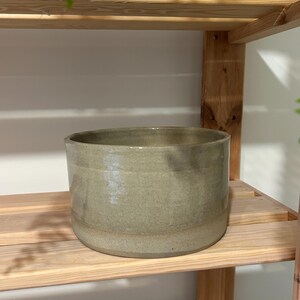May include: A cylindrical ceramic planter with a smooth, glossy finish in a muted sage green color. The vessel features a wide, open top and a flat base, showcasing a consistent, solid tone throughout its exterior surface and a simple, clean design.