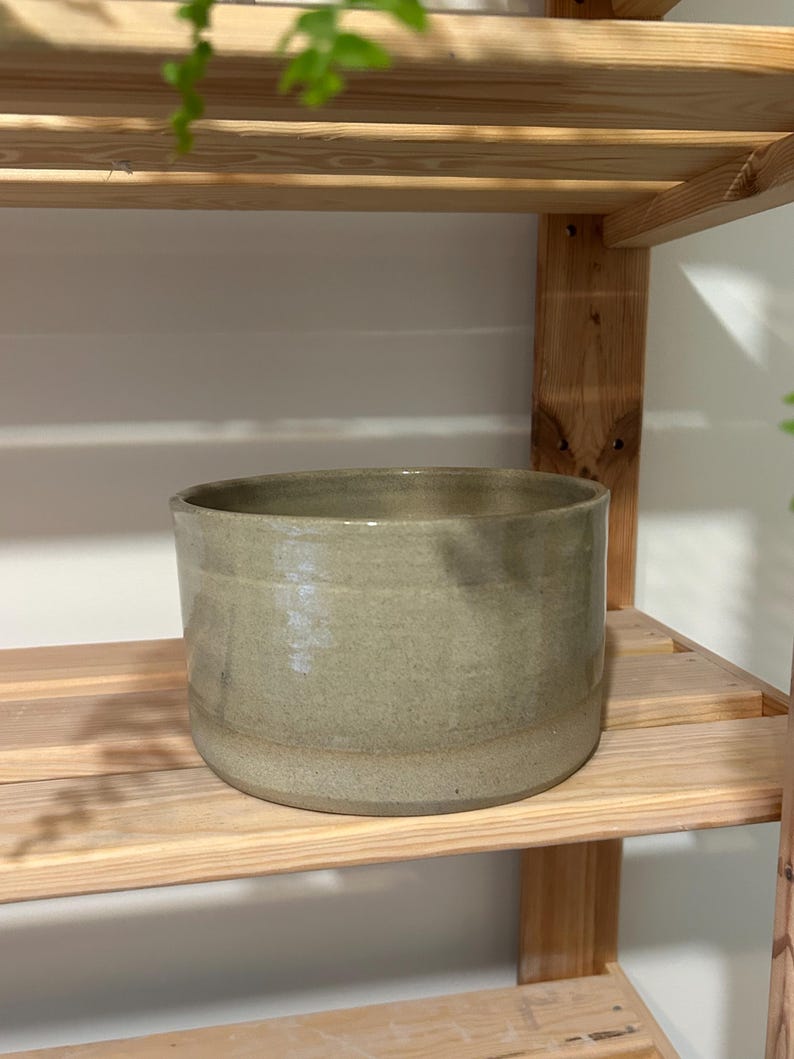 May include: A cylindrical ceramic planter with a smooth, glossy finish in a muted sage green color. The vessel features a wide, open top and a flat base, showcasing a consistent, solid tone throughout its exterior surface and a simple, clean design.