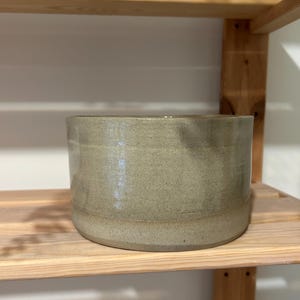 May include: A cylindrical ceramic bowl with a smooth, light sage green glaze and a subtle speckled texture throughout the surface. The vessel features a flat base and straight, vertical walls, showcasing a consistent finish and a simple, functional design.