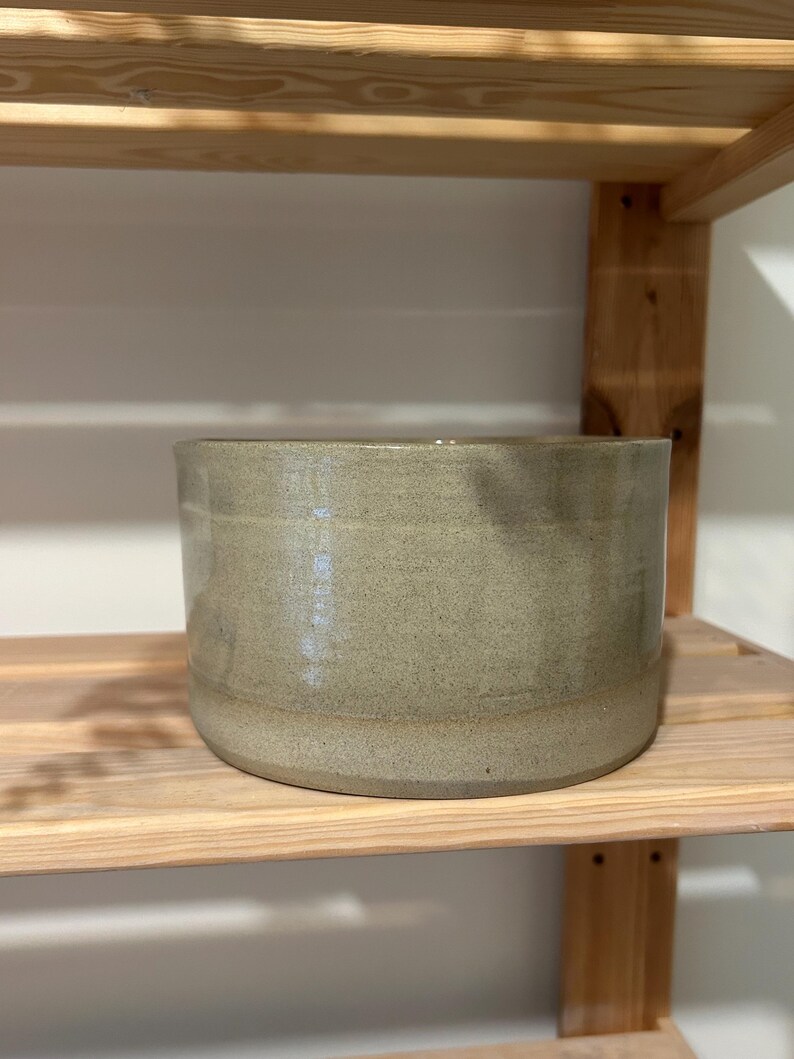 May include: A cylindrical ceramic bowl with a smooth, light sage green glaze and a subtle speckled texture throughout the surface. The vessel features a flat base and straight, vertical walls, showcasing a consistent finish and a simple, functional design.