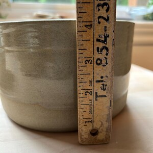 May include: A cylindrical ceramic bowl with a smooth, light sage green glaze. A vintage wooden ruler is placed vertically against the side of the bowl, showing a height measurement of approximately 5 inches. The ruler features black text.