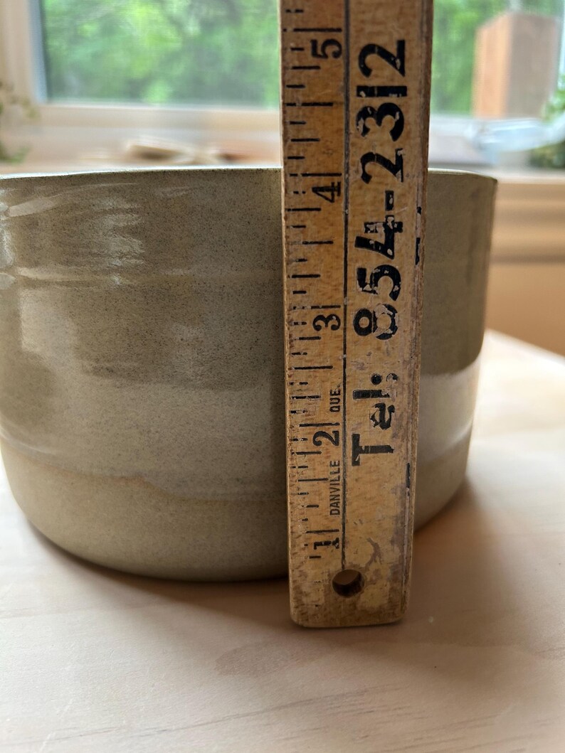 May include: A cylindrical ceramic bowl with a smooth, light sage green glaze. A vintage wooden ruler is placed vertically against the side of the bowl, showing a height measurement of approximately 5 inches. The ruler features black text.