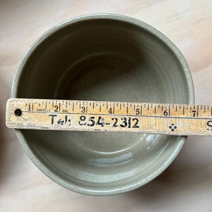 May include: A round, light olive-green ceramic bowl with a smooth interior finish. A wooden ruler is placed across the top, showing a diameter of approximately 6.5 inches. The ruler features the text "Danville Que." and "Tel: 854-2312".