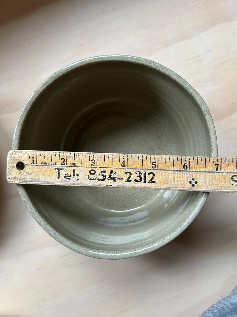 May include: A round, light olive-green ceramic bowl with a smooth interior finish. A wooden ruler is placed across the top, showing a diameter of approximately 6.5 inches. The ruler features the text "Danville Que." and "Tel: 854-2312".
