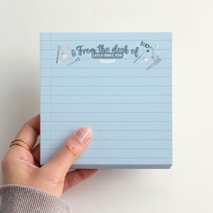May include: A light blue notepad with horizontal lines, held by a hand. The notepad has the text "From the desk of" and dental-themed illustrations. The name "LAYLA JONES, RDH" is also printed on the notepad.
