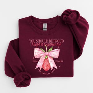 May include: A maroon sweatshirt with the text "YOU SHOULD BE PROUD I kept it together for nine months TOO CUTE TO BE ALONE" and a watercolor strawberry design with a pink bow.