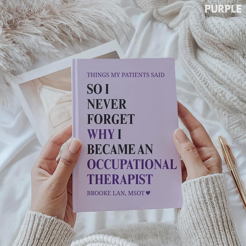 May include: A lilac notebook with the text "THINGS MY PATIENTS SAID SO I NEVER FORGET WHY I BECAME AN OCCUPATIONAL THERAPIST" is held open. A gold pen rests on the white surface. The word "PURPLE" is in the top right corner.