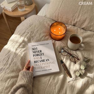 May include: A relaxing setup with a lit orange candle in a glass jar, a white mug with tea, and a book titled "So I Never Forget Why I Became an Occupational Therapist." Also present are eyeglasses and a pen, all on a beige bed.
