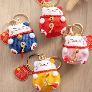Peut inclure: Porte-clés chats porte-bonheur crochetés en rouge, bleu, jaune et rose. Chaque chat a un visage blanc, une clochette dorée et un pompon rouge. Les porte-clés ont des anneaux dorés et des breloques rouges en forme de cœur.