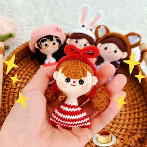 Puede incluir: Cuatro muñecas de crochet, cada una con un atuendo y un peinado diferentes. Las muñecas están sentadas en una cesta de mimbre. La muñeca del centro lleva un vestido a rayas rojas y blancas y un lazo rojo.