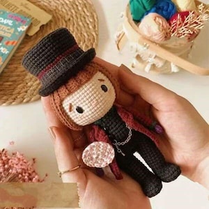 Può includere: Una bambola all'uncinetto con i capelli castani, che indossa un cappello a cilindro nero, una giacca rosso e nera e pantaloni neri. La bambola tiene un oggetto all'uncinetto rosa e bianco nella sua mano destra.