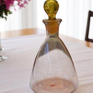 Könnte beinhalten: Eine klare Glaskaraffe mit breiter Basis und schmalem Hals. Die Karaffe hat einen gelben Glasstopfen mit abgerundeter Oberseite. Die Karaffe steht auf einer weißen Tischdecke.