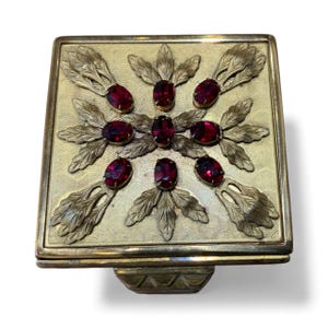 May include: A square, gold-toned compact with a decorative floral design. The compact features eight oval, deep red gemstones and stylized leaf accents. The compact has a textured surface and a small, decorative base.