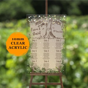 May include: A clear acrylic wedding seating chart with a hand-drawn illustration of a tree with two hearts. The chart lists guest names by table number. The text reads "Erica & David" and "17 April 2023".