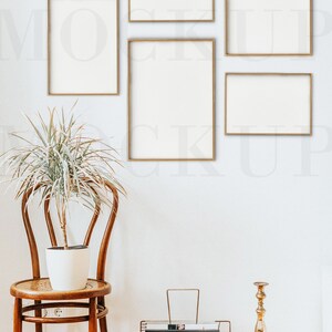 May include: Five gold picture frames of various sizes hang on a white wall. A potted plant sits on a wooden chair in front of the frames. A gold magazine rack with a book titled "Around the Homes" sits on the wooden floor. A gold candlestick stands next to the magazine rack.