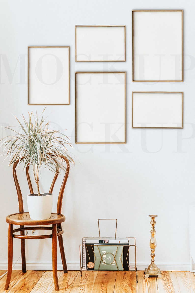 May include: Five gold picture frames of various sizes hang on a white wall. A potted plant sits on a wooden chair in front of the frames. A gold magazine rack with a book titled "Around the Homes" sits on the wooden floor. A gold candlestick stands next to the magazine rack.
