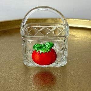 May include: A small glass basket with a clear diamond pattern and a red tomato design. The basket has a handle and is sitting on a gold tray.