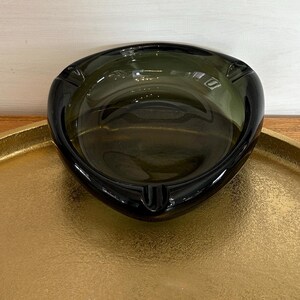 May include: A dark green glass ashtray sits on a gold metal tray. The ashtray has a rounded, triangular shape with a slightly raised rim.