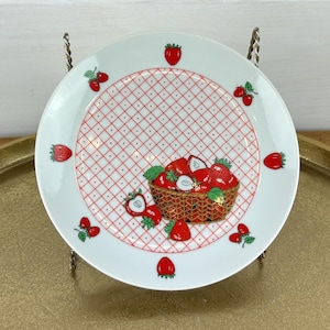 May include: A white ceramic plate with a red and white checkered pattern and strawberry illustrations. The plate features a central image of a basket overflowing with strawberries. Additional strawberries are placed around the rim.
